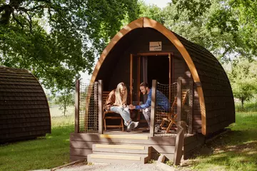 A man shows a woman his phone on the porch of their wooden cabin surrounded by green foliage