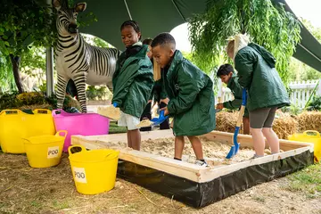 Children wearing zookeeper coats shovelling through sand in a sand pit