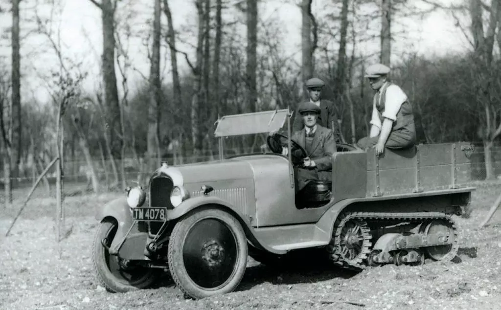 A Citroen half-tracked vehical being driven down a slope at Whipsnade in 1929. The driver as accompanied by two other men. There is woodland in the background. 