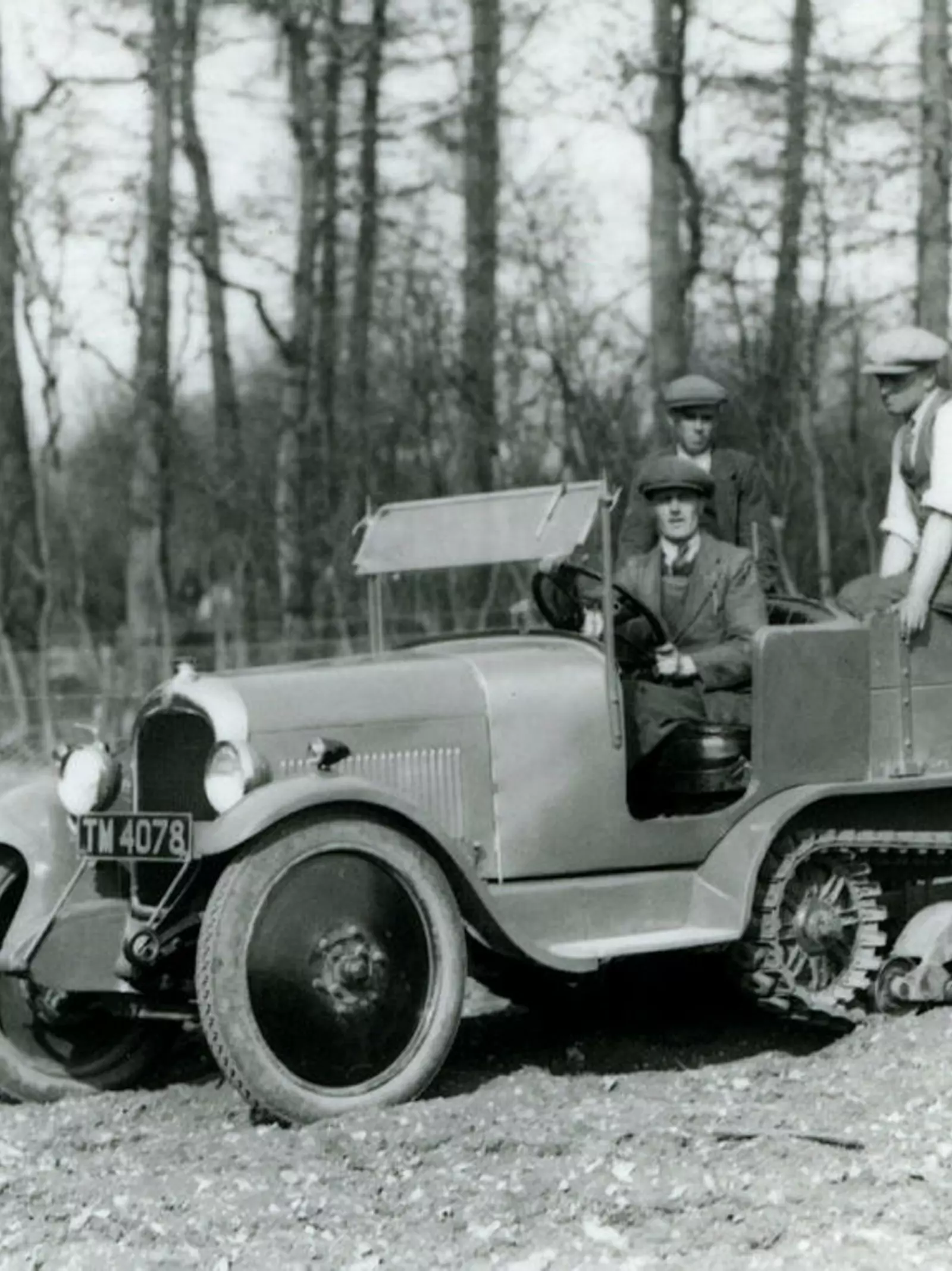 A Citroen half-tracked vehical being driven down a slope at Whipsnade in 1929. The driver as accompanied by two other men. There is woodland in the background. 