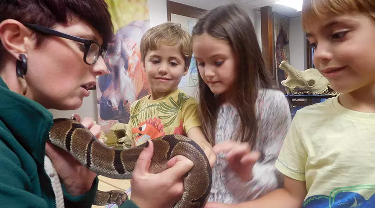 Three children participating in the Zoo Explorers course look at a snake held by a member of the Whipsnade Zoo staff