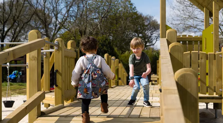 A young boy and a female toddler walk towards each other on a wooden play platform