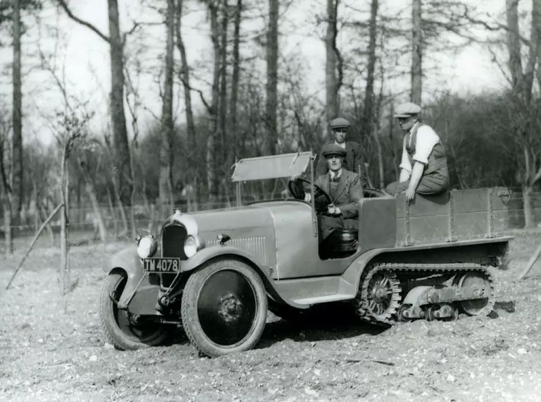A Citroen half-tracked vehical being driven down a slope at Whipsnade in 1929. The driver as accompanied by two other men. There is woodland in the background. 