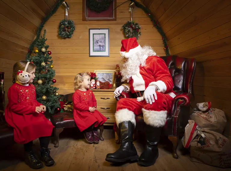 Santa sits in his wooden lodge grotto across from two young, caucasian girls wearing matching red dresses and smiling up at him