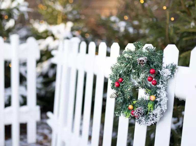 A Christmas wreath hangs from a white picket fence on the left hand side of the frame, while in the background is snow-laden foliage and twinkly lights