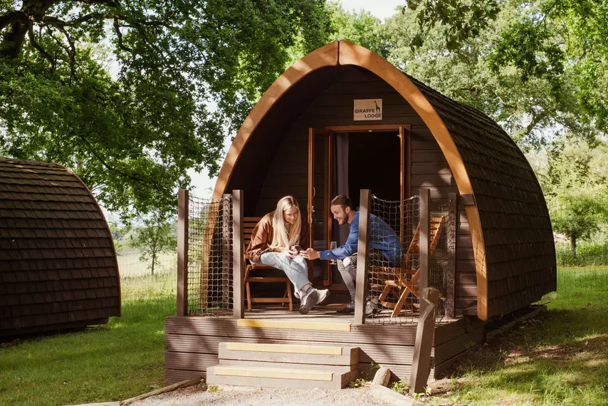 A man shows a woman his phone on the porch of their wooden cabin surrounded by green foliage